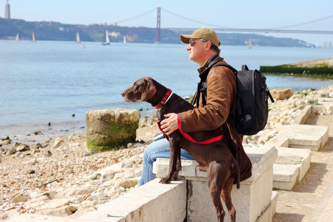 A man and his dog by the sea, in the distance is the 25 de Abril Bridge which looks remarkably like the Golden Gate Bridge.